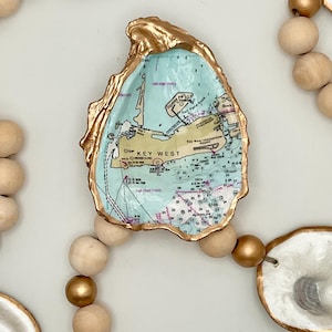 May include: A decorative garland featuring natural oyster shells with gold-painted edges and a decoupage map of Key West on the center shell. The shells are strung together with alternating natural wood beads and metallic gold-painted wooden beads.