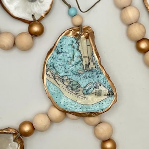 May include: A coastal-themed garland featuring oyster shells with gold-painted edges, each displaying a map of Sanibel Island. The garland includes natural wooden beads and gold-painted beads, strung together with twine and a blue bead.