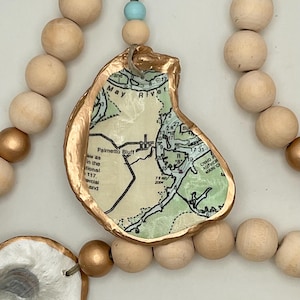 May include: A decorative beaded garland featuring oyster shells with map designs. The shells have a gold-colored trim and are interspersed with natural wooden beads and gold-painted beads. A small blue bead accents the design.