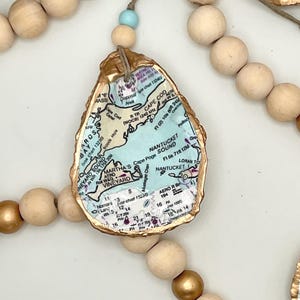 May include: A decorative garland featuring natural wooden beads, metallic gold beads, and a light blue bead. The garland includes oyster shells with gold-painted edges, each featuring a decoupage map of Cape Cod, Martha's Vineyard, and Nantucket.