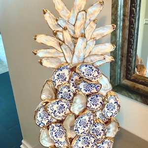 May include: A decorative pineapple sculpture constructed from numerous oyster shells. The top leaves consist of iridescent white shells, while the base features shells decorated with a blue and white floral pattern, all with gold-painted edges.