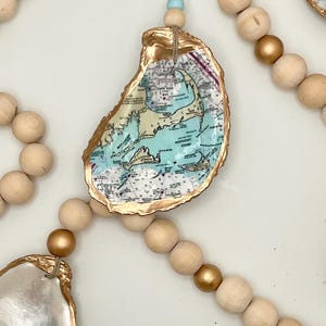 May include: A decorative garland featuring natural wooden beads in matte and metallic gold finishes, interspersed with oyster shells. One central shell displays a nautical map of Cape Cod and Nantucket Sound, with edges painted in a metallic gold finish.