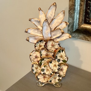 May include: A decorative pineapple sculpture crafted from individual oyster shells. The top leaves are white with gold-painted edges, while the base features shells decoupaged with white magnolia flower patterns and gold-trimmed borders on a stand.