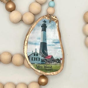 May include: A decorative oyster shell pendant featuring a lighthouse image with the text "Tybee Island, Georgia." The shell has a gold-painted edge and is strung with wooden beads and a blue accent bead. A coastal-themed ornament.