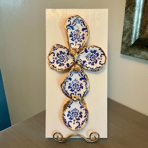 May include: A wall-mounted cross decoration featuring four oyster shells arranged in a cross shape on a white rectangular wooden board. Each shell is painted with a blue and white floral pattern and finished with a gold-painted edge.