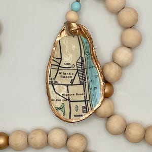 May include: A decorative oyster shell ornament featuring a map of Atlantic Beach and Neptune Beach with gold-painted edges, suspended from a string of natural wooden beads and metallic gold beads, accented by a single small light blue bead.