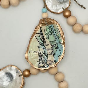 May include: A decorative garland featuring oyster shells with gold-painted edges and a map of St. Augustine, Florida. The garland includes wooden beads in natural and gold tones, and a light blue bead.