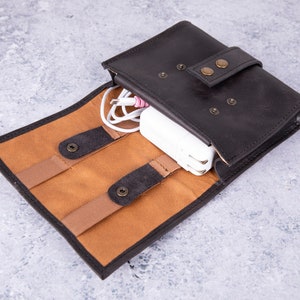 May include: A brown leather travel case with a tan interior and elastic straps to hold cords and a small white power adapter. The case has a flap with a snap closure and a brown leather strap with a brass button.