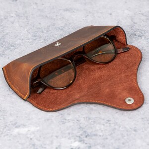 May include: A brown leather glasses case with a snap closure and a brown suede lining. The case is open and a pair of glasses with brown frames and clear lenses are inside.