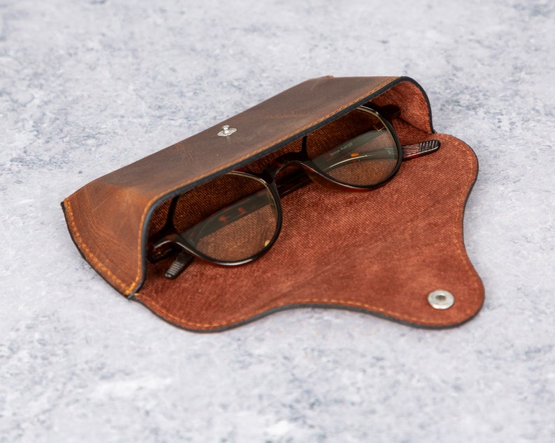 May include: A brown leather glasses case with a snap closure and a brown suede lining. The case is open and a pair of glasses with brown frames and clear lenses are inside.