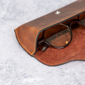 May include: A brown leather glasses case with a pair of glasses inside. The case has a flap that closes with a snap.