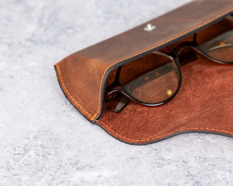 May include: A brown leather glasses case with a pair of glasses inside. The case has a flap that closes with a snap.