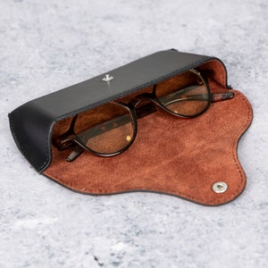 May include: A black leather glasses case with a brown suede interior. The case is open and a pair of brown-tinted glasses are inside.