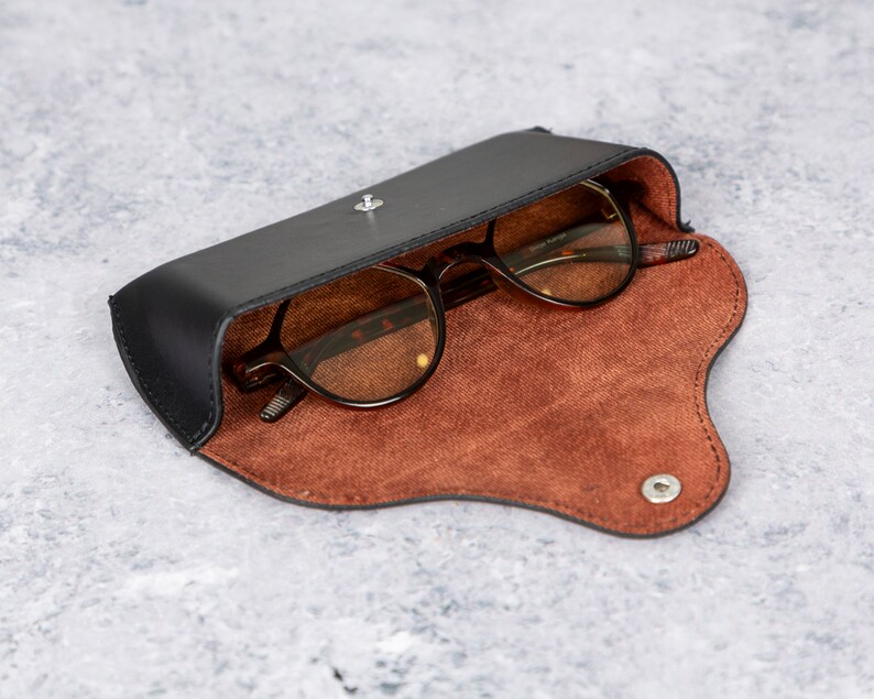 May include: A black leather glasses case with a brown suede interior. The case is open and a pair of brown-tinted glasses are inside.