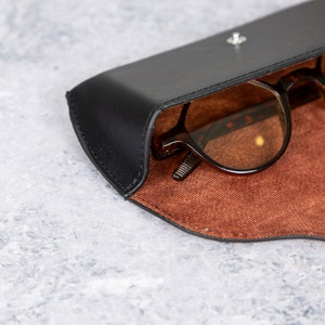 May include: A black leather glasses case with a brown suede interior. The case is open and a pair of brown glasses is inside.