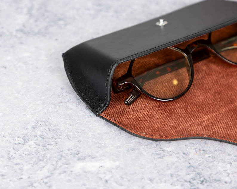May include: A black leather glasses case with a brown suede interior. The case is open and a pair of brown glasses is inside.