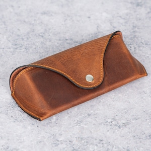 May include: A brown leather glasses case with a snap closure. The case has a stitched seam around the edge and a silver button.