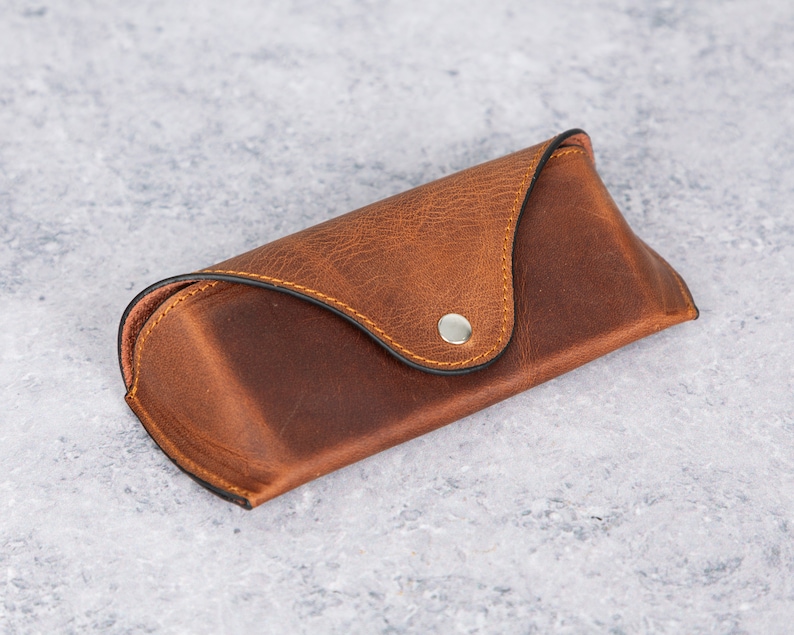 May include: A brown leather glasses case with a snap closure. The case has a stitched seam around the edge and a silver button.