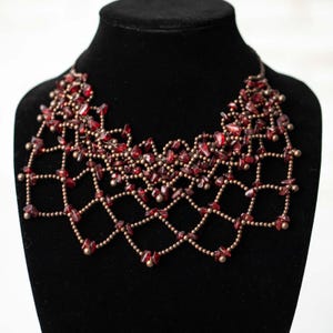 May include: A statement necklace featuring a lattice design constructed from small metallic copper-toned beads. The structure is embellished with numerous irregular, deep red translucent stone chips woven throughout the grid-like pattern.