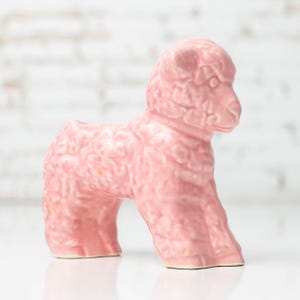 May include: A small, light pink ceramic figurine shaped like a lamb. The surface features a textured, curly wool pattern covering the body and head. The lamb is standing in a profile position, facing toward the right, with a smooth, glossy finish.