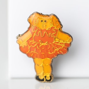 May include: A metal enamel pin shaped like a doll with a round head and curly hair. The doll wears a bright orange dress with a ruffled skirt and a bow at the waist. The figure has yellow legs and small black shoes, outlined in silver metal.