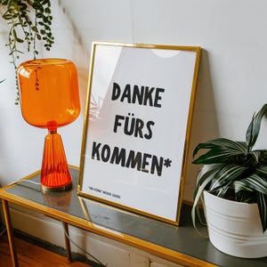 Könnte beinhalten: Ein goldgerahmter Druck mit dem deutschen Text "DANKE FÜRS KOMMEN" in Schwarz. Eine orangefarbene Glaslampe und eine Topfpflanze stehen auf einem Tisch. Die Lampe hat einen zylindrischen Schirm und einen konischen Fuß.