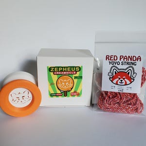 May include: A white and orange yoyo with a cat face cutout, a white box with a Zepheus Creamsicle label featuring a cat graphic, and a clear plastic bag containing five pieces of red and white Red Panda Yoyo String with a cat logo label.