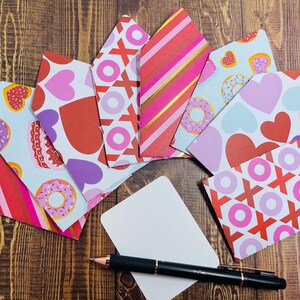 May include: A set of colorful envelopes with different patterns, including hearts, donuts, and stripes. The envelopes are arranged in a fan shape on a wooden surface. A blank card and a black pen are also included.
