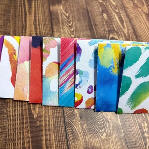 May include: A row of ten colorful paper squares with abstract watercolor designs. The squares are arranged in a line from left to right, with the colors gradually transitioning from purple to green.