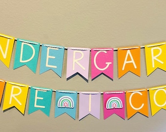 May include: A colorful banner with the words "KINDERGARTEN HERE COME" strung across two lines. Each pennant is a different color, including pink, orange, yellow, and teal, with white lettering. Rainbow stickers are on some pennants.