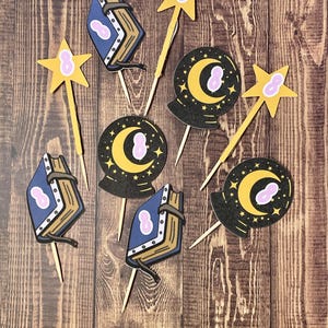 May include: A set of eight cupcake toppers featuring a whimsical magical theme. The toppers include a blue book with a gold clasp, a crescent moon with stars, and a yellow star with a pink center.