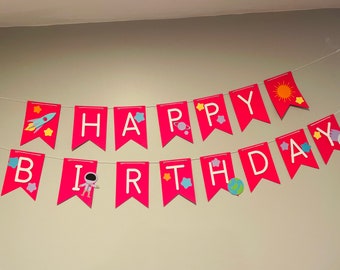 May include: A pink and white banner with the words "Happy Birthday" in white letters. The banner has a space theme with stars, planets, rockets, and astronauts.