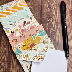 May include: A stack of colorful patterned envelopes with a blank white card and a black pen on a wooden surface.