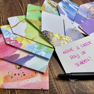 May include: A stack of colorful envelopes with watercolor designs and gold accents. The top envelope has a handwritten message that says "Have a great day at school!"
