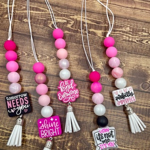 May include: Five pink and white silicone bead necklaces with different charms. The charms include the phrases "tomorrow needs you", "shine bright", "be kind to everyone", "perfectly imperfect", and "listen it's not me it's you". Each necklace has a white tassel.
