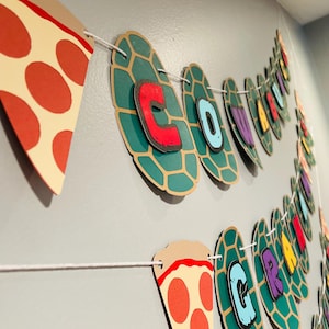 May include: A Teenage Mutant Ninja Turtles themed party banner with green turtle shells and pizza slices. The banner spells out "Cowabunga!" in colorful letters.