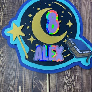 May include: A layered cake topper with a celestial theme. It features a gold crescent moon, stars, and a wand. The number "8" and the name "ALEX" are in holographic, iridescent colors. A small book is on the right side.