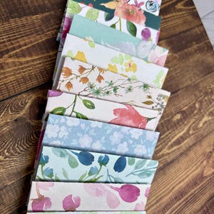 May include: A stack of 12 floral patterned envelopes in various colors, including pink, blue, green, and yellow. The envelopes are arranged in a vertical stack, with the top envelope being a light purple with a floral pattern.