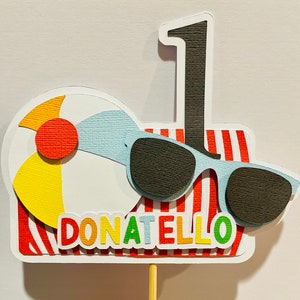 May include: A red, white, and blue striped background with a white beach ball, black sunglasses, and a black number one. The text "DONATELLO" is written in rainbow colors below the sunglasses.
