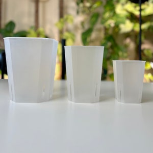 May include: Three translucent white, hexagonal plant pots of varying sizes arranged in a row. Each pot features a tapered shape with vertical drainage slits located near the base, providing a clear view of the structure and design of the containers.