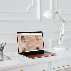 Puede incluir: Un espacio de trabajo moderno con un port&aacute;til de oro rosa que muestra una p&aacute;gina web, un calendario peque&ntilde;o, una planta suculenta en una maceta gris y una l&aacute;mpara de escritorio blanca. El escritorio es blanco con tiradores dorados, contra una pared blanca.