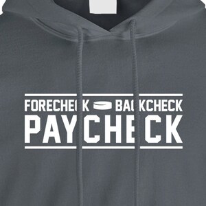 May include: A dark gray hooded sweatshirt with the text "FORECHECK BACKCHECK PAYCHECK" in white lettering. A hockey puck icon is between the words "FORECHECK" and "BACKCHECK".