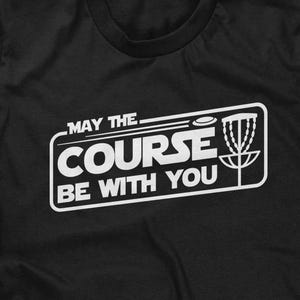 May include: Black t-shirt with a white graphic that reads "MAY THE COURSE BE WITH YOU." The design includes a disc golf basket and a disc. The shirt is a crew neck style.