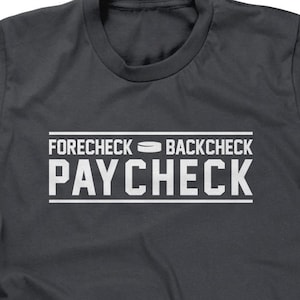 May include: A dark gray t-shirt with white text that reads "Forecheck Backcheck Paycheck" with a hockey puck symbol between the words "Forecheck" and "Backcheck".
