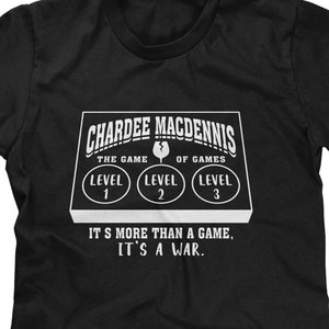 May include: Black t-shirt with a white graphic of a game board with the text "CHARDEE MACDENNIS THE GAME OF GAMES" and three buttons labeled "LEVEL 1", "LEVEL 2", and "LEVEL 3". The text "IT'S MORE THAN A GAME, IT'S A WAR." is below the game board.