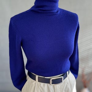 Peut inclure: Un pull à col roulé bleu roi à manches longues. Il est associé à un pantalon beige et une ceinture bleu foncé avec une boucle argentée. L'ensemble est présenté sur un mannequin, mettant en valeur un style classique et polyvalent.