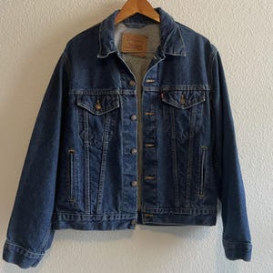 May include: A dark blue denim jacket with a classic design. The jacket features a button-up front, two chest pockets, and two side pockets. The jacket has a collar and long sleeves. The Levi's brand tag is visible on the inside.