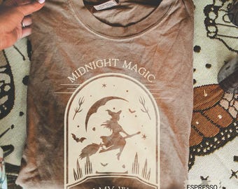 May include: A brown crew neck t-shirt featuring a cream-colored graphic of a witch flying on a broomstick under a crescent moon. The text reads MIDNIGHT MAGIC ON MY WAY. The shirt is labeled as espresso color and has a standard short-sleeve design.