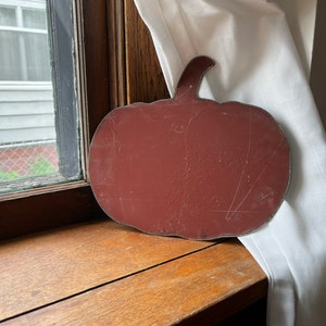 May include: A rusty red metal pumpkin cutout with a stem. The pumpkin is sitting on a wooden surface next to a white curtain.