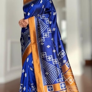 May include: A royal blue sari with a geometric white pattern and a gold border. The sari is draped over the body. The fabric has a sheen. The sari is a traditional garment.